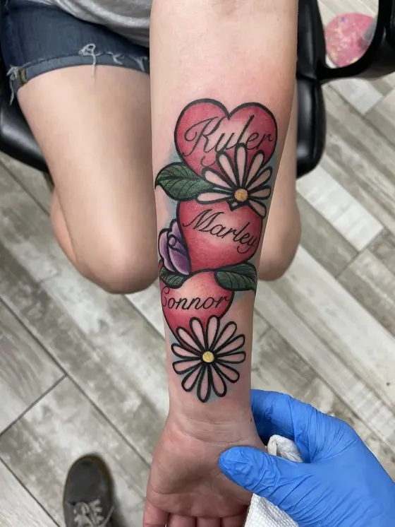 Color traditional style tattoo of hearts, daisies and family names across a client's forearm, tattooed at TBA Tattoos
