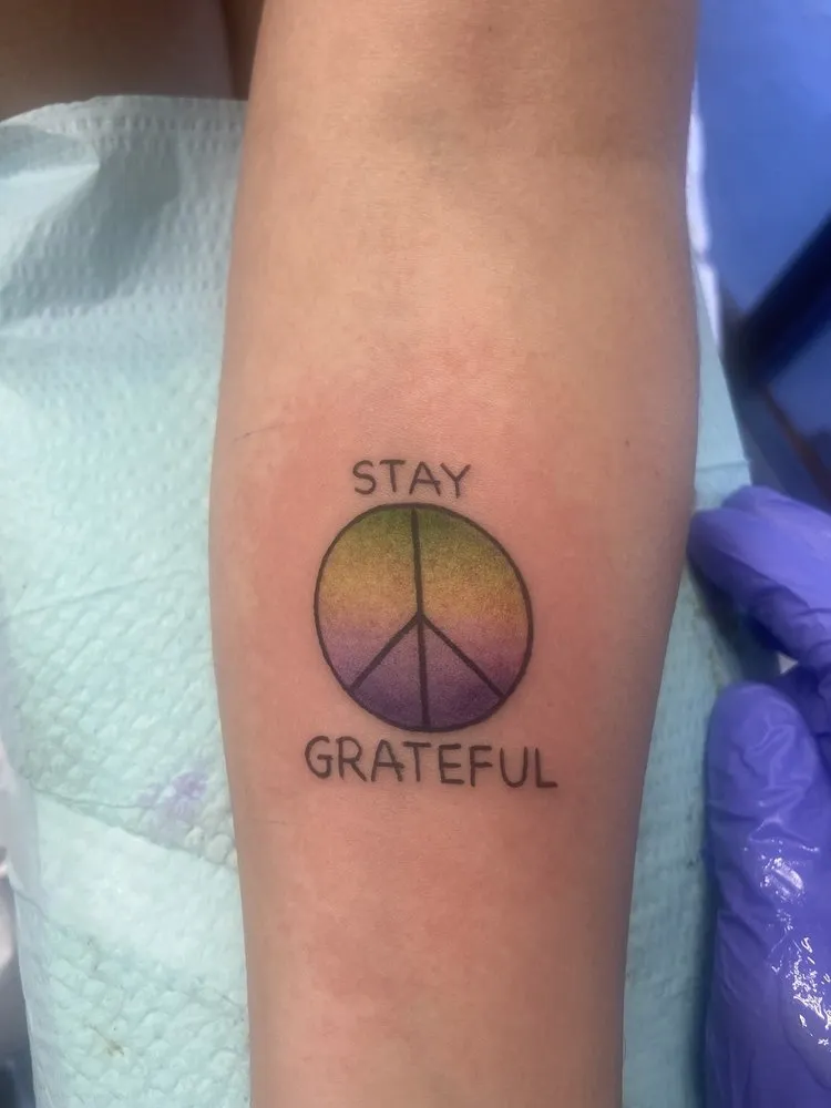 Small color tattoo of a hand flashing a peace sign with the words Stay Grateful, tattooed at TBA Tattoos in West Palm Beach